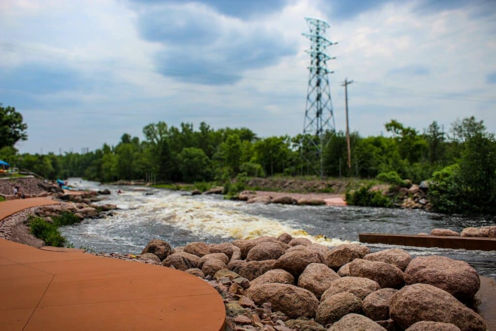 A river by a pathway, surrounded by trees and rocks, what you will see at the Wausau Whitewater Park.